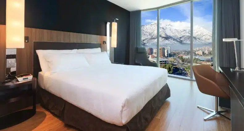Quarto do Icon Hotel, em Santiago, com cama de casal no centro do quarto do lado esquerdo do quarto janelas panorâmicas com vista das montanhas, para representar hotéis com vista para as Cordilheiras dos Andes em Santiago.