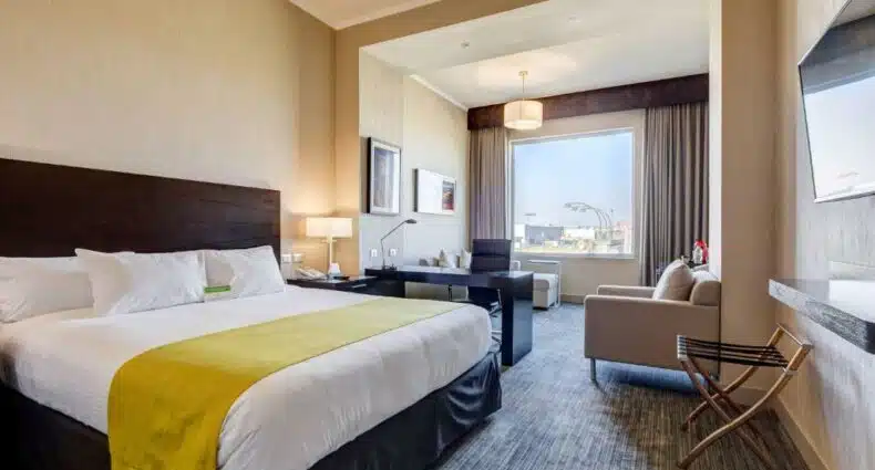 Quarto do La Quinta by Wyndham Santiago Aeropuerto, com cama de casal do lado direito, do lado esquerdo mesa com cadeira e uma poltrona bege. Representa hotéis perto do aeroporto de Santiago.