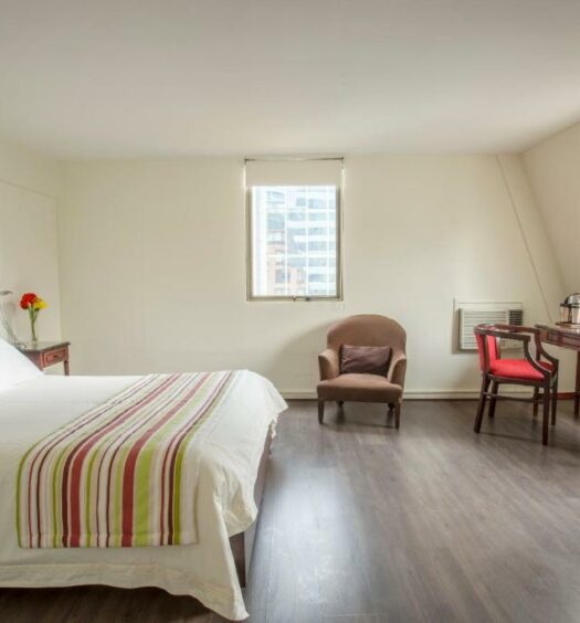 Quarto do MR Express (ex Hotel Neruda Express), com cama de casal no centro do quarto do lado esquerdo, uma poltrona do lado esquerdo e uma mesa de madeira com cadeira em frente a cama. Representa hotéis baratos em Santiago.