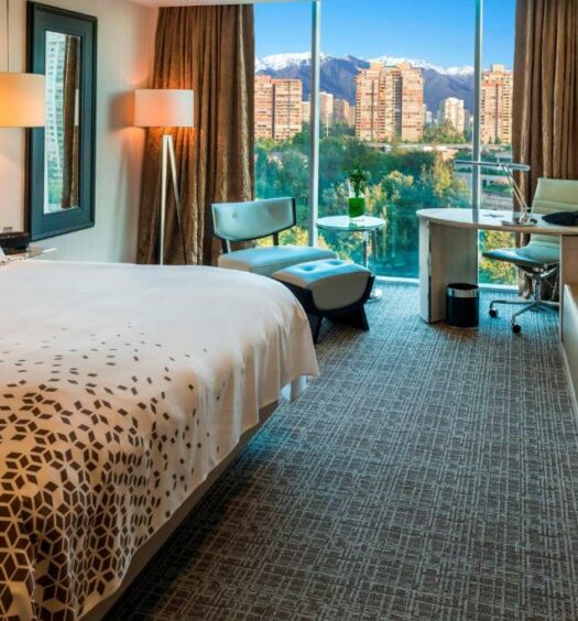 Quarto do Renaissance Santiago Hotel com cama de casal do lado esquerdo e em frente a cama cômoda de madeira e do lado esquerdo do quarto poltrona e uma mesa de trabalho. Representa hotéis românticos em Santiago.