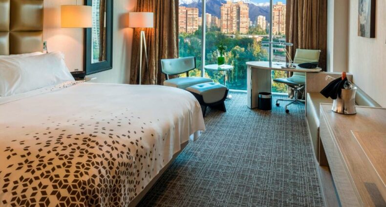 Quarto do Renaissance Santiago Hotel com cama de casal do lado esquerdo e em frente a cama cômoda de madeira e do lado esquerdo do quarto poltrona e uma mesa de trabalho. Representa hotéis românticos em Santiago.