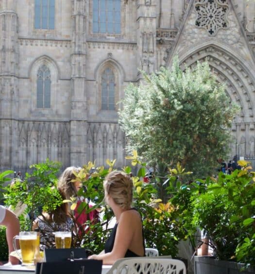 Casal sentado em mesa no Colón Hotel Barcelona, uma das recomendações de hotéis românticos em Barcelona. Eles olham para a Catedral de Barcelona que está atrás, e há algumas árvores ao redor. A foto foi tirada durante o dia.