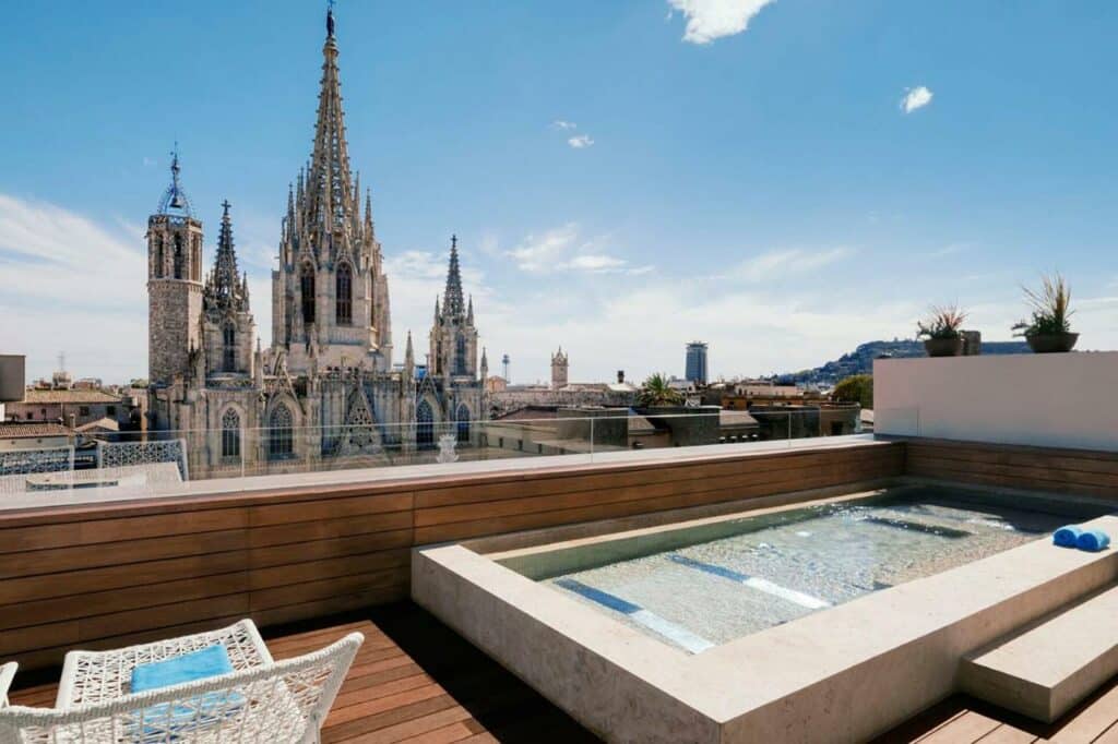 Área da piscina do Colón Hotel Barcelona, uma das recomendações de hotéis românticos em Barcelona. A vista direta é para a Catedral de Barcelona. Há uma espreguiçadeira branca ao lado da pequena piscina no deque.
