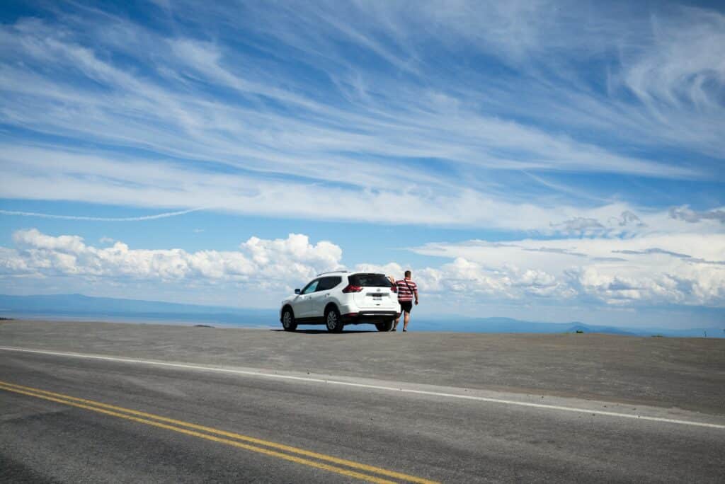 Carro branco parado em estrada de Crater Lake, em Oregon, nos EUA, com pessoa parada ao lado direito, em dia de céu claro repleto de nuvens.