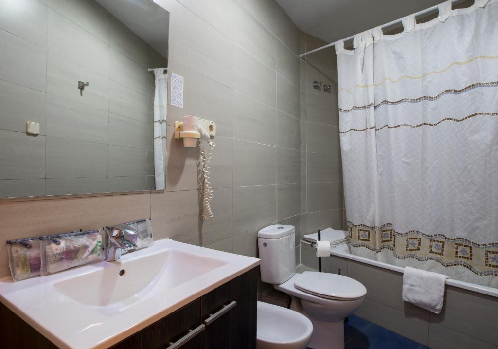 Banheiro do Cuatro Naciones. Há uma banheira atrás de uma cortina de banho e ao lado da privada branca. A pia tem amenidades de banho em cima e um espalho atrás. O secador de cabelo está fixado na parede ao lado do espelho.