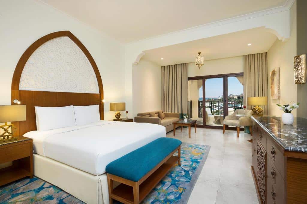 Quarto do DoubleTree by Hilton Ras Al Khaimah com uma varanda com vista para a praia, uma ante sala com sofá e poltronas, um quarto com uma cama de casal e um móvel com gavetas, há alguns itens de decoração em tons de dourado