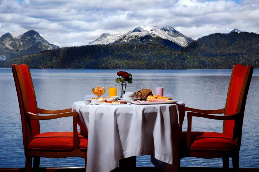 Mesa redonda com toalha branca e café da manhã em cima, com duas cadeiras vermelhas (uma de cada lado) e uma vista para o lago e montanhas ao fundo