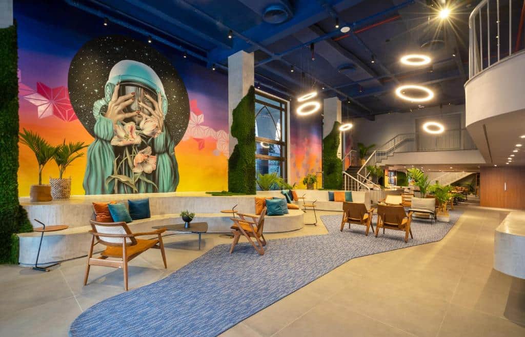 Recepção do hotel, colorido, com tapete azul e cadeiras de madeira. Na parede um banco com almofadas para as pessoas sentarem. Na parede da esquerda uma arte colorida com um capacete de astronalta.