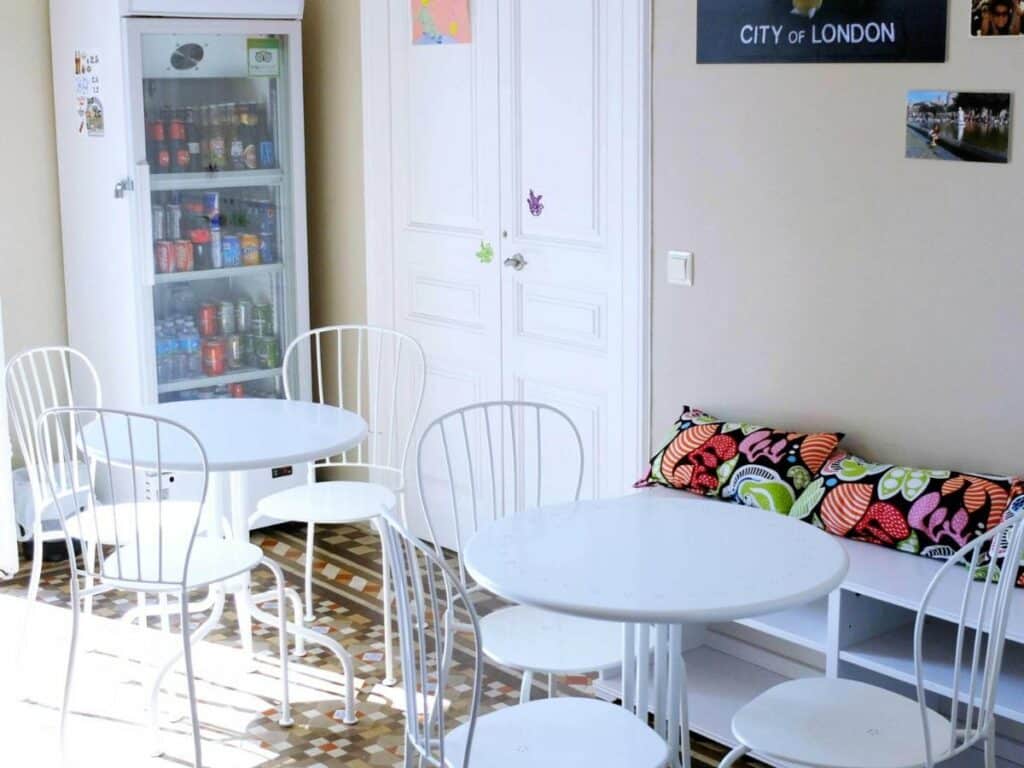 Área comum do Fabrizzio's Petit, uma das recomendações de hostels em Barcelona. Duas mesas brancas com cadeiras estão no lugar, e algumas almofadas coloridas estão em um móvel ao lado da mesa mais próxima. Ao fundo, uma geladeira com bebidas está ao lado de uma porta.
