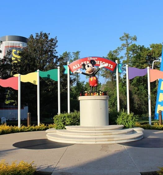 fachada com o nome do hotel atrás de uma estátua de braços abertos do Mickey com bandeirinhas coloridas de chegada dos lados e cones de torcida gigantes atrás no Disney's All-Star Sports Resort, um dos resorts da Disney