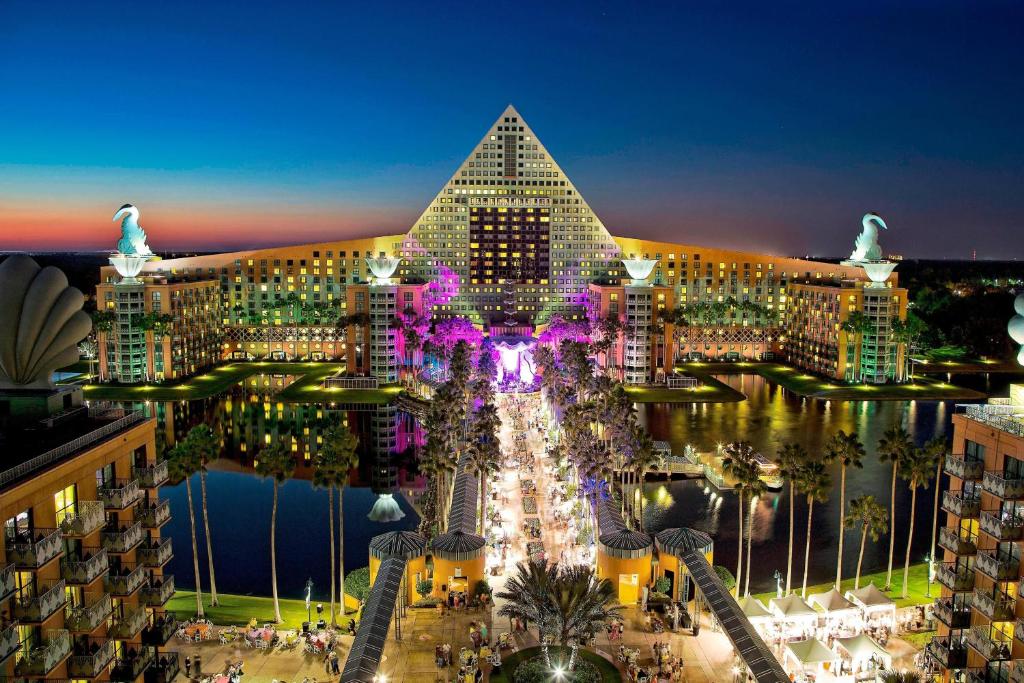 fachada do Walt Disney World Swan Reserve, um dos hotéis da Disney em Orlando, com muitas luzes, uma passarela acima do lago e uma estrutura em forma de pirâmide com cisnes, tudo bem rebuscado