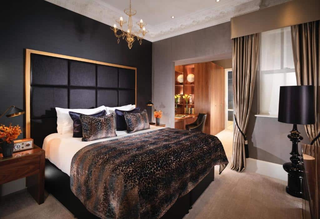 Quarto do Flemings Mayfair com uma cama de casal com diversas almofadas, uma pequeno lustre no teto, chão de carpete, uma janela com cortinas e um abajur de chão