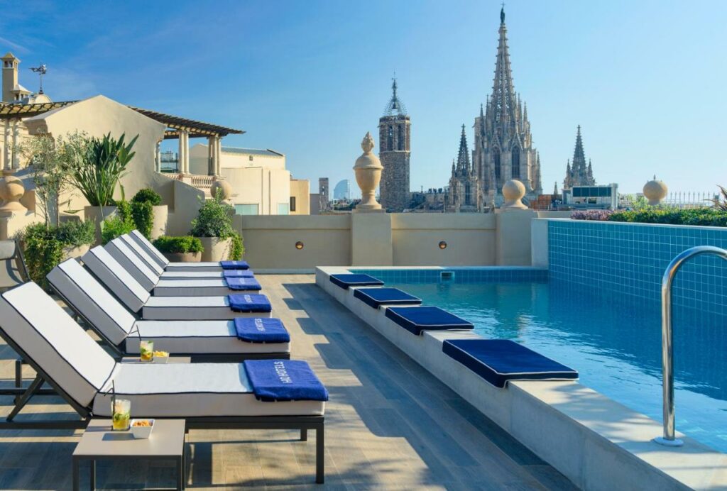 Área da piscina do H10 Madison, uma das recomendações de hotéis no Bairro Gótico em Barcelona. Com vista para a Catedral de Barcelona, a área tem espreguiçadeiras brancas intercaladas com mesinhas. Elas encaram a piscina que tem escadas e almofadas na beirada.