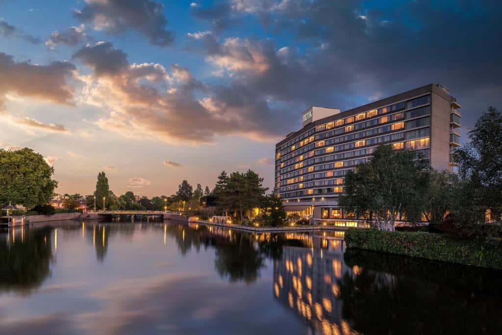 Vista do Hilton Amsterdam, um dos hotéis de luxo, durante um início de noite com o céu começando a escurecer, com um lago a frente refletindo as luzes do prédio e, no lado direito, o prédio do hotel com várias janelas com luzes acesas e algumas arvores em volta