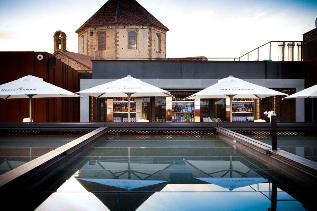 Piscina do Hotel España Ramblas, uma das recomendações de hotéis nas Ramblas em Barcelona. Guarda-sóis brancos estão abertos ao longo da extensão da piscina que reflete a imagem deles. Ao fundo está o hotel.