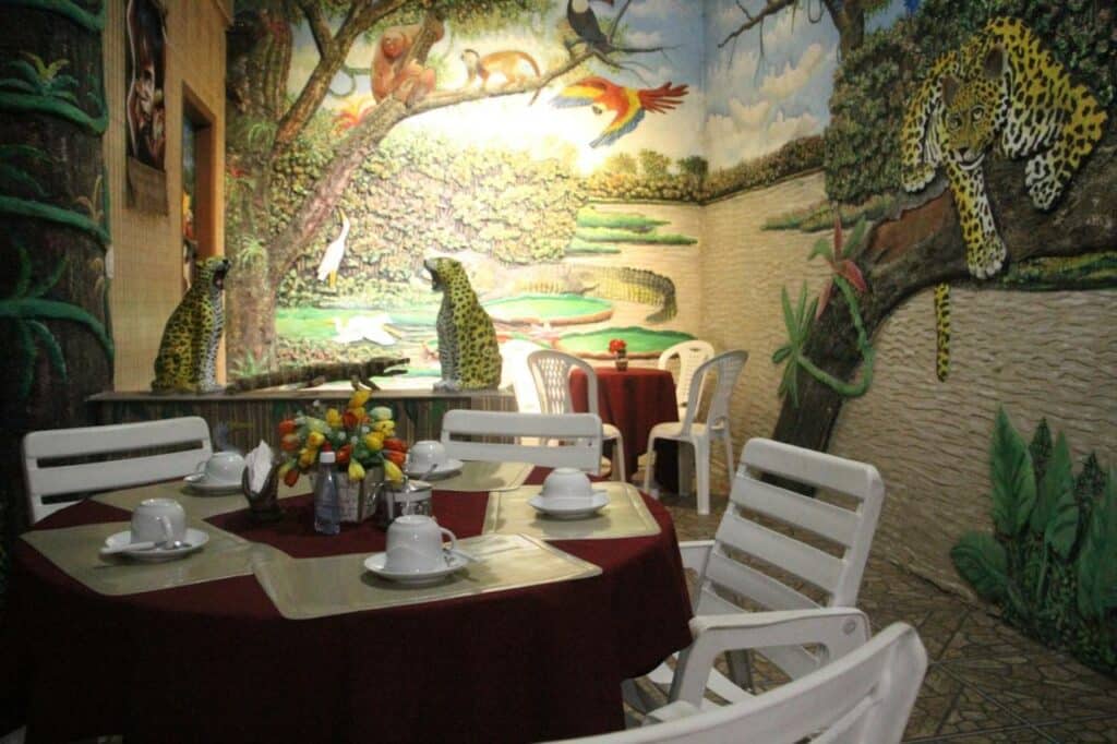 Área de refeição toda decorada com desenhos de animais e floresta. Há mesas com cadeiras, xícaras e flores.