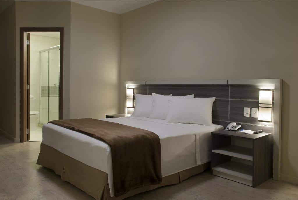 Uma cama de casal no meio, luzes dos dois lados acesas, uma cômoda dos dois lados, e no canto esquerdo a porta do banheiro aberta.