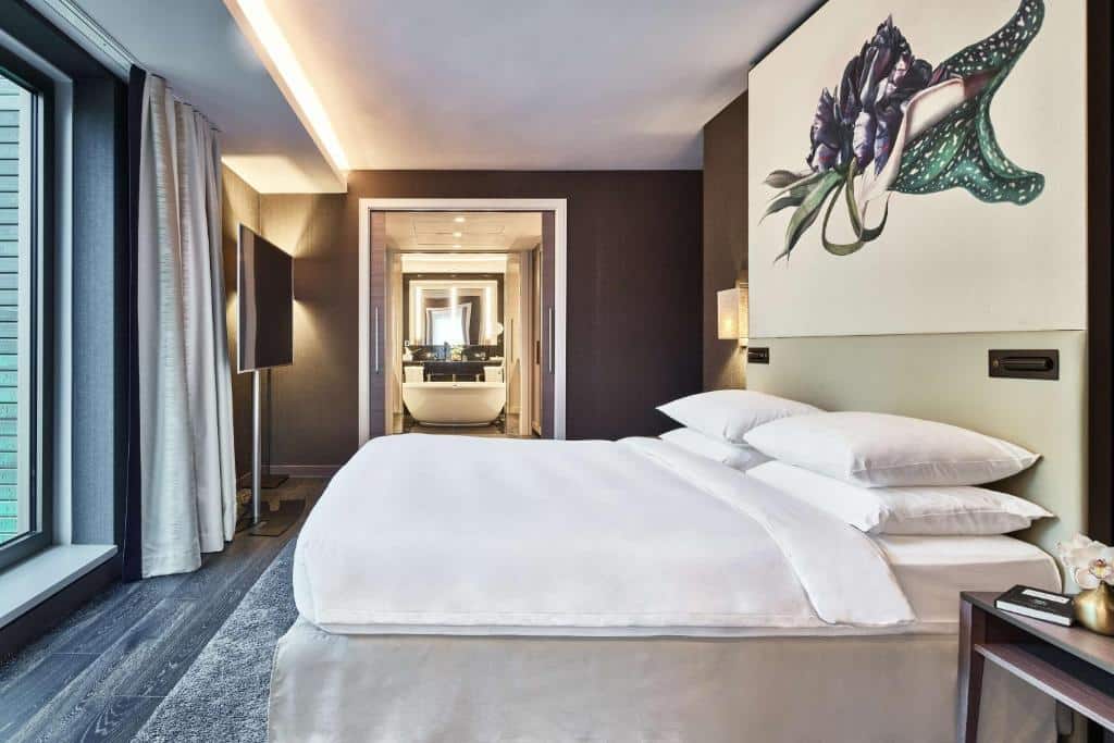 Interior da Suíte Presidencial do Hyatt Regency Amsterdam, de 126 m², com uma cama de casal e um quadro com desenho de flor verde e rocha, e ao fundo a porta do banheiro está aberta, aparecendo a banheira de hidromassagem