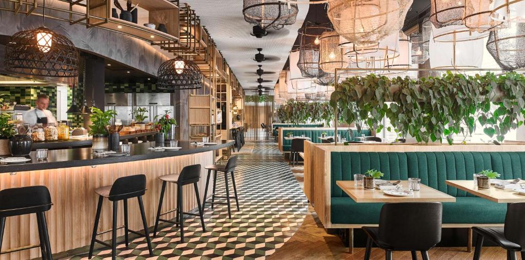 Restaurante do Hyatt Regency Amsterdam, um dos hotéis de luxo, com piso preto e branco, no lado direito, tem mesas e cadeiras com estofado verde e, no lado esquerdo, tem um balcão com a cozinha atrás e uma pessoa. O local é todo decorado com luminárias e folhas verdes