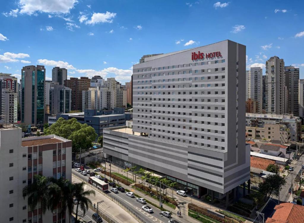 Vista aérea de São Paulo, e o hotel ibis São Paulo Ibirapuera está em destaque em meio a outros prédios. A foto foi tirada de dia, está ensolarado com algumas nuvens no céu. Vários carros passam na rua.