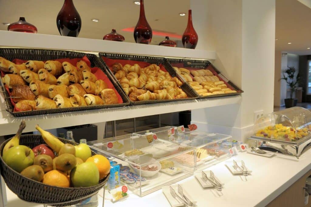 Café da manhã do Ibis Cornella. Pães, frutas e frios estão dispostos no balcão com pegadores de metal em frente.