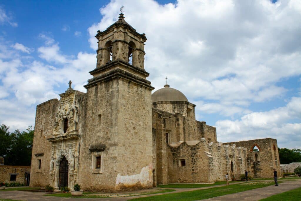 Uma construção de tijolos e pedras bem antiga e desgastada que faz parte do Parque Histórico Nacional das Missões de San Antonio, há uma torre em destaque e, mais ao fundo, uma abóboda