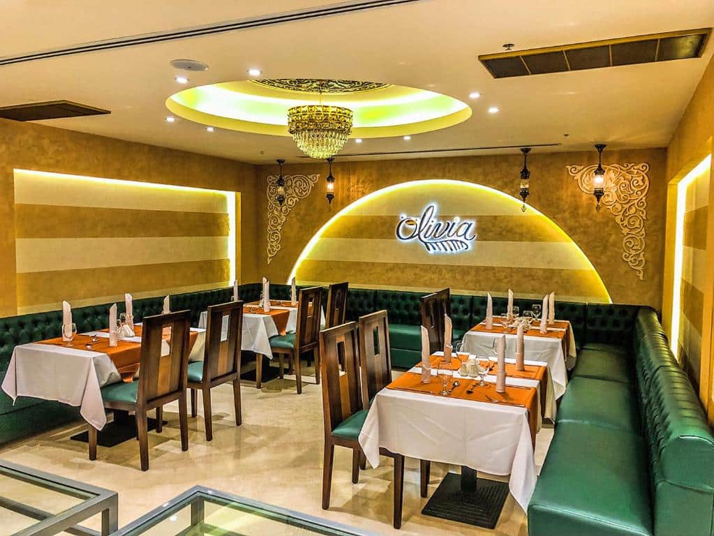 Salão para refeições no Mangrove Hotel com alguns detalhes em dourado, um lustre redondo, mesas quadradas, sofás verdes e cadeiras de madeira