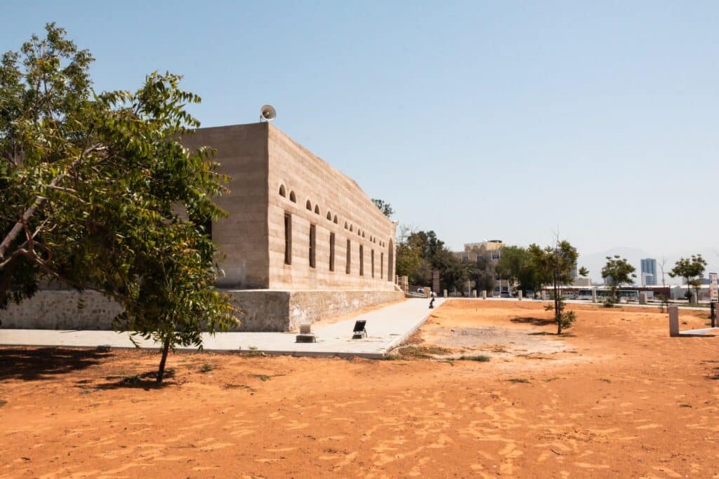 Uma construção histórica de tijolos bege com diversas janelas, no arredores há areia alaranjada e uma árvore, o local é conhecido como Mohammed Bin Salem Mosque, para representar o que fazer em Ras Al Khaimah