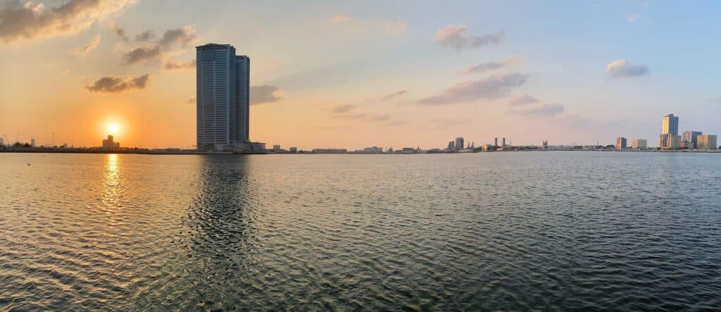 Vista do mar em Ras Al Khaimah com prédios ao fundo do horizonte e o sol de pondo do lado esquerdo da foto