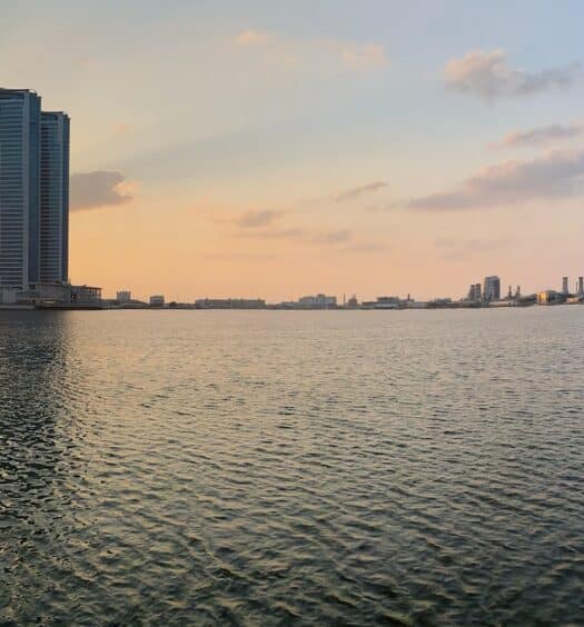 Vista do mar em Ras Al Khaimah com prédios ao fundo do horizonte e o sol de pondo do lado esquerdo da foto