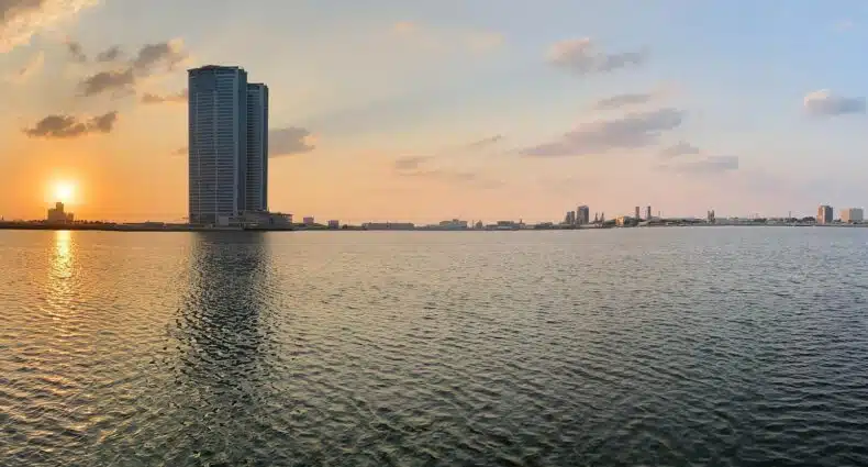 Vista do mar em Ras Al Khaimah com prédios ao fundo do horizonte e o sol de pondo do lado esquerdo da foto