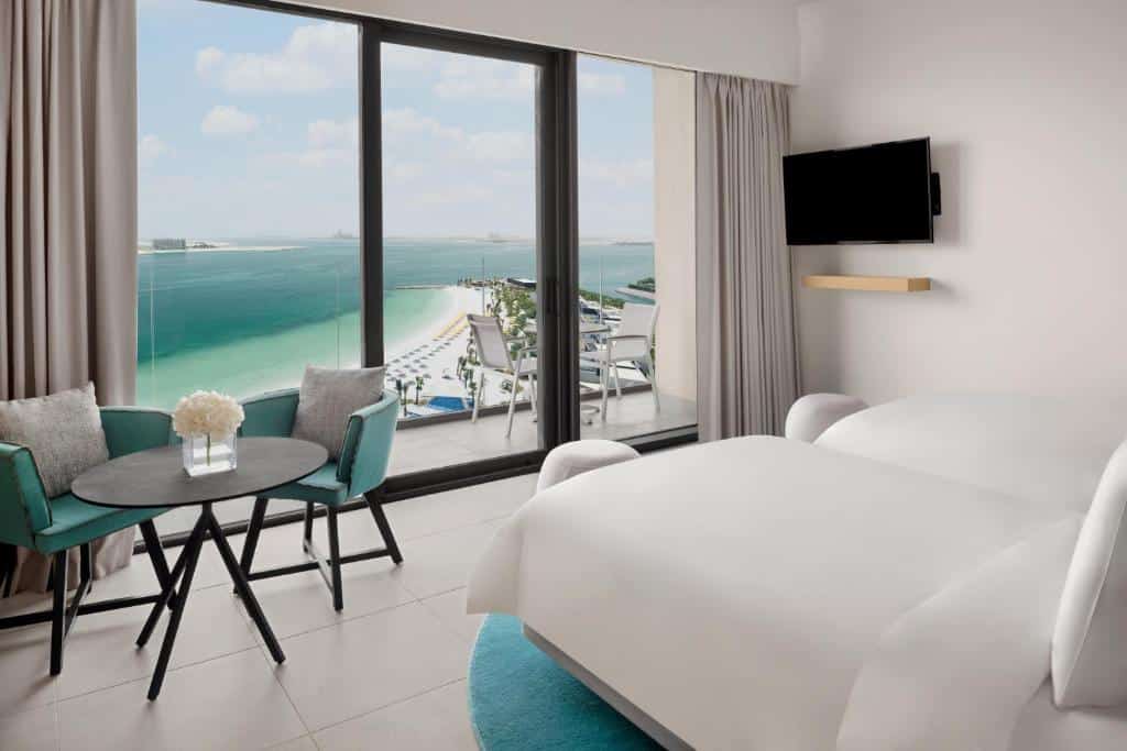 Quarto do Mövenpick Resort Al Marjan Island com uma varanda ampla com vista para o mar, duas camas de solteiro, uma mesinha com duas poltronas, uma televisão e um tapete azul claro
