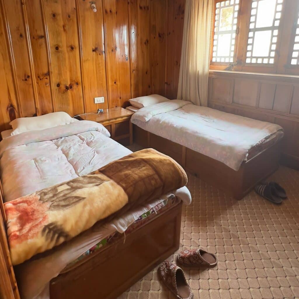Quarto com duas camas de solteiro, uma escrivaninha ao meio, uma janela no lado esquerdo e uma cabeceira de madeira rústica