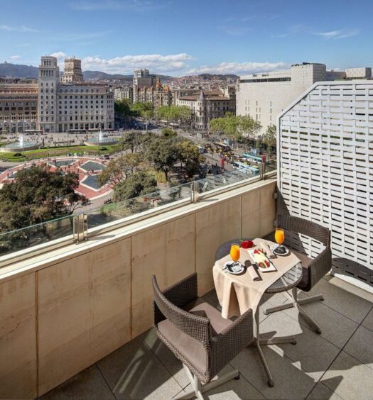 Sacada de um quarto do Olivia Plaza Barcelona, uma das recomendações de hotéis no Bairro Gótico em Barcelona. Uma mesa com café da manhã e duas cadeiras está encostada na balaustrada que tem vista para a Plaça de Catalunya. A foto foi tirada durante o dia e o sol ilumina o local.
