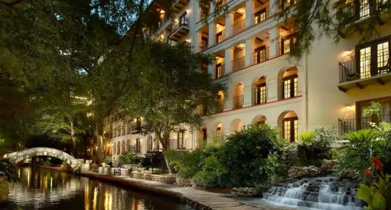 Prédio do Omni La Mansion del Rio virado para o River Walk com muitas árvores ao redor e uma pequena fonte do lado direito, para representar hotéis em San Antonio