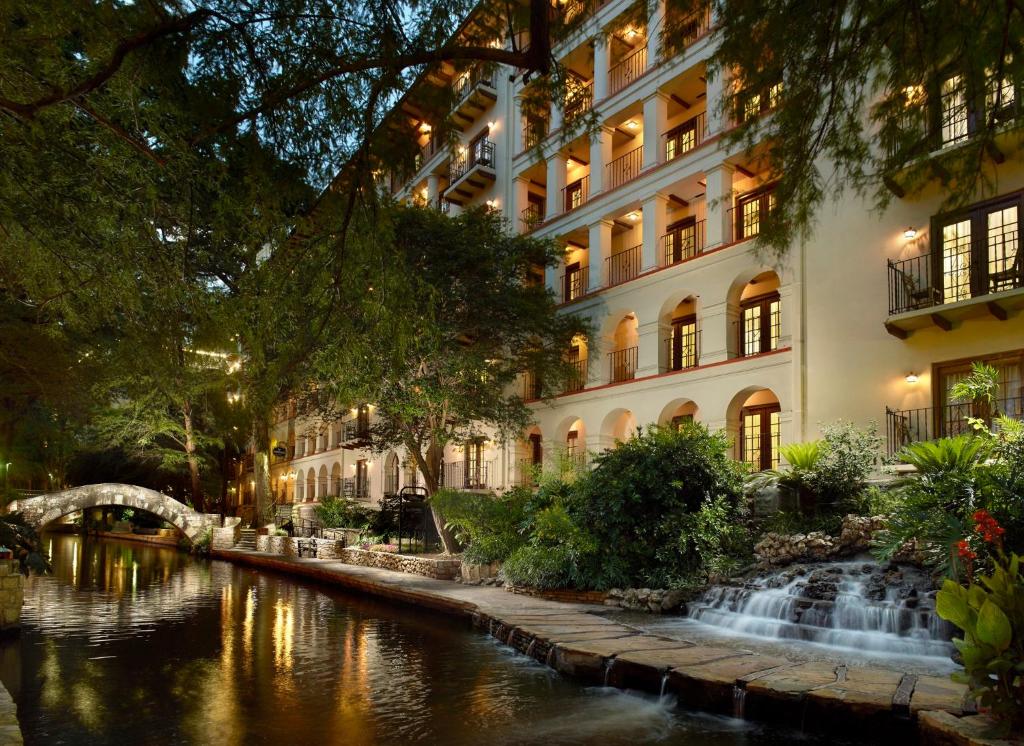 Prédio do Omni La Mansion del Rio virado para o River Walk com muitas árvores ao redor e uma pequena fonte do lado direito, para representar hotéis em San Antonio