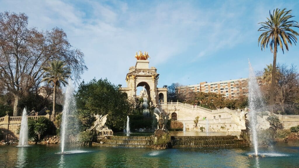 Lago do Parc de la Ciutadella para ilustrar o post sobre aluguel de carro em Barcelona. Chafarizes sopram água para cima ao redor de uma construção à frente. Há árvores ao redor e a foto foi tirada de dia, com um céu azul acima. - Foto: VENUS MAJOR via Unsplash