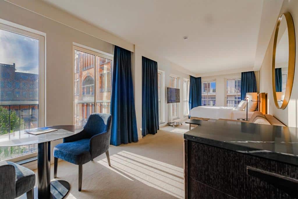 Estúdio Premium do Pestana Amsterdam Riverside, de 33 m², com janelas amplas no quarto todo com vista panorâmica para prédios históricos e algumas cortinas azuis, cama de casal, sofá, espelho redondo e um móvel preto ao lado.