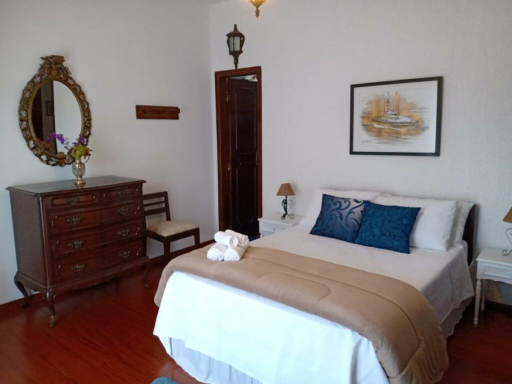 Quarto da Pousada Canto da Paz, com cama ao centro com lençóis brancos e travesseiros azuis, mesa de cabeceira e estante com gavetas e espelho acima ao canto.