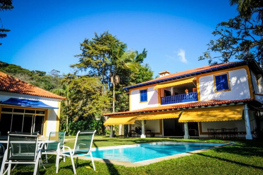 Fachada da Pousada Vila Brasil em um dia de céu azul com sol, a pousada ao fundo rodeado de vegetação e uma piscina em primeiro plano com uma mesa e cadeiras próximas.