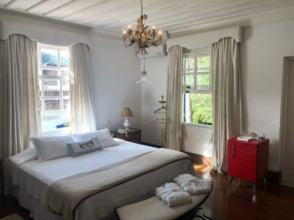 Quarto moderno da Pousada Vila Brasil com uma cama de casal, uma janela com vista para a vegetação, todos os móveis são de madeira, um frigobar vermelho de canto e uma mesa de cabeceira com abajour em cima.