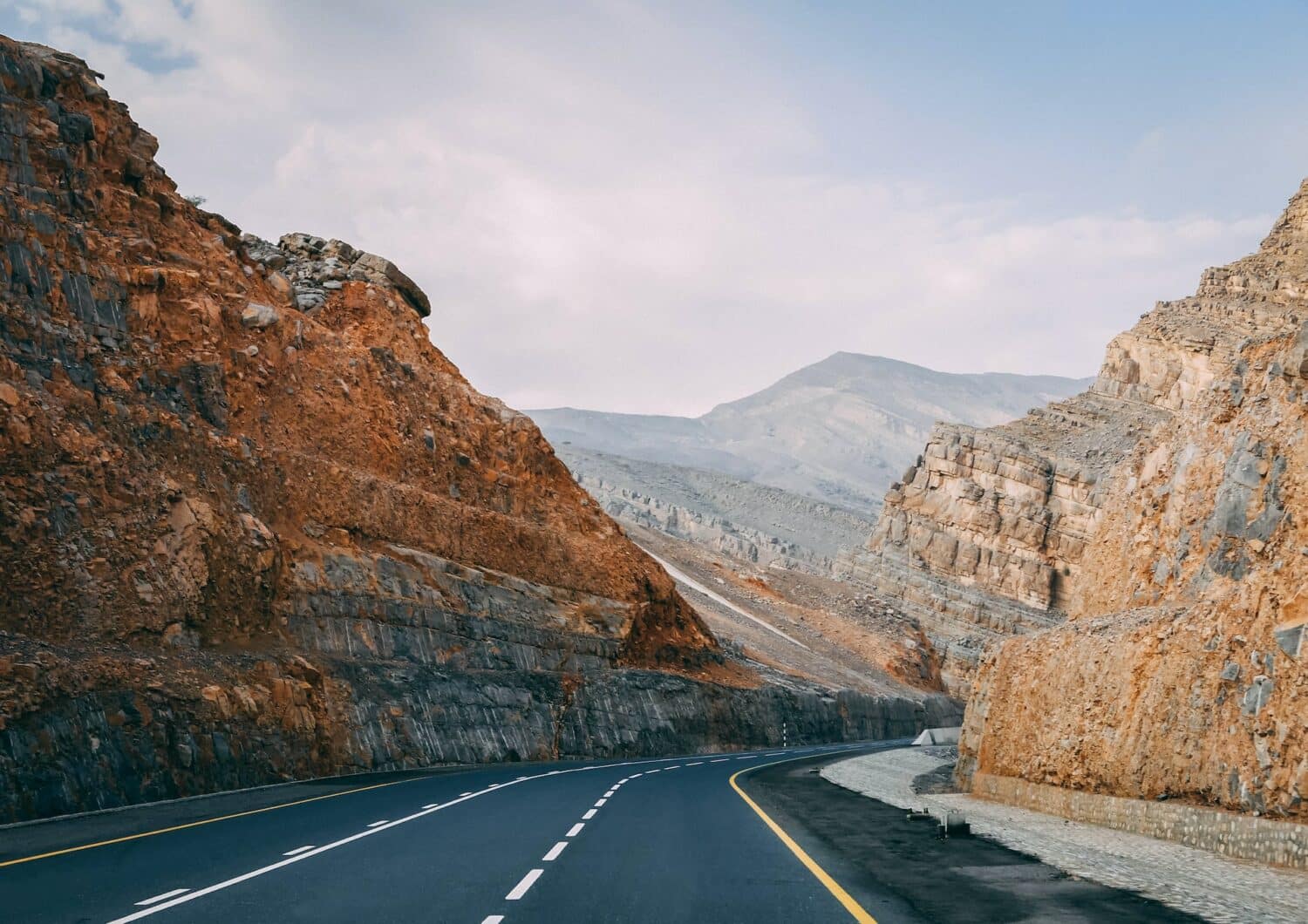 Uma estrada cortando montanhas rochosas em tons de cinza, laranja e bege, esse local é conhecido como Jebel Jais em Ras Al Khaimah