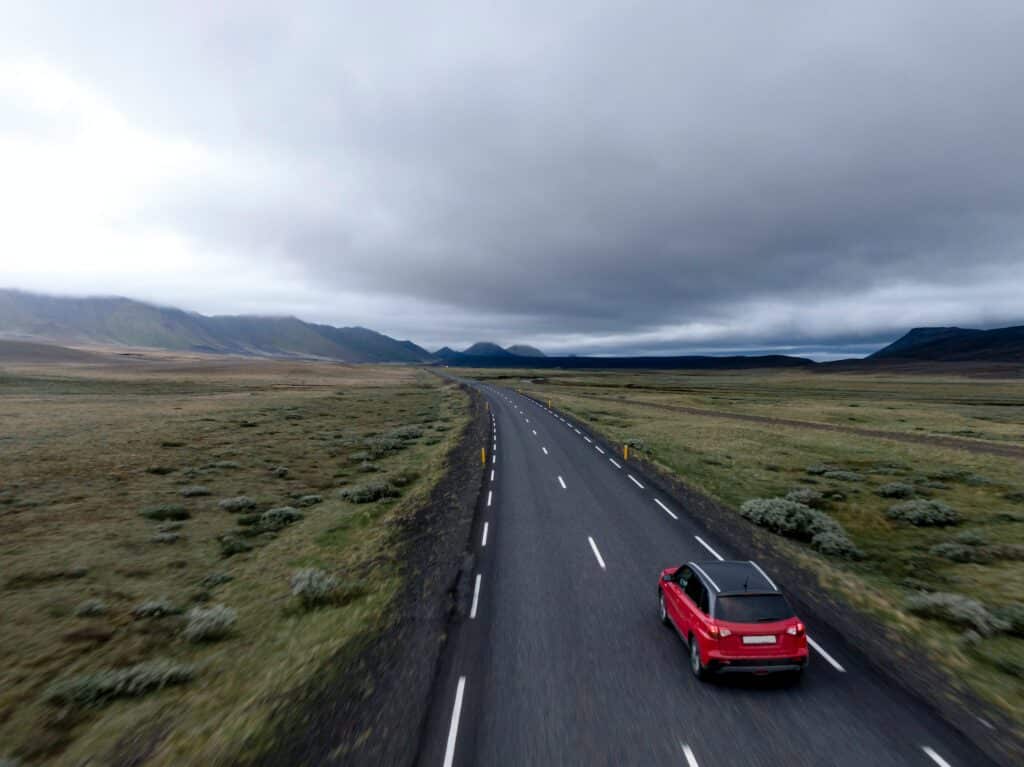 Carro vermelho com teto preto em estrada com campos gramados em ambos os lados, cadeira de montanhas ao fundo, pela esquerda, e nuvens densas ocupando o céu.