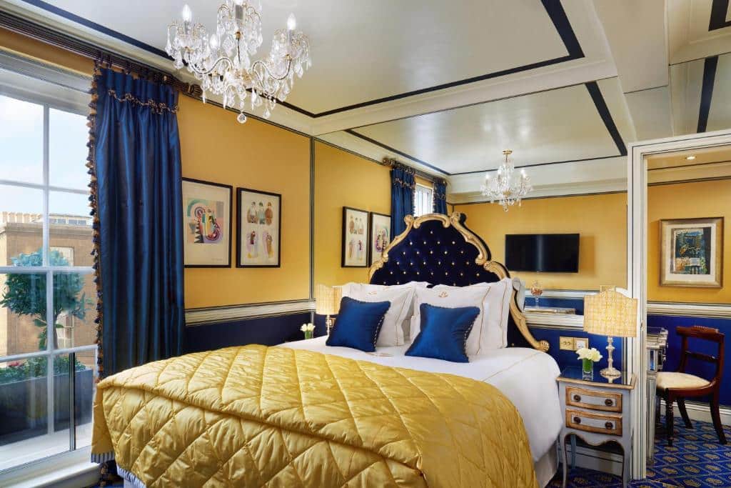 Quarto do Rubens At The Palace com tudo decorado em tons de dourado e azul, há uma janela com cortinas, uma lustre, uma amplo espelho na cabeceira e duas mesinhas de cabeceira com abajures, para representar hotéis em Londres para brasileiros