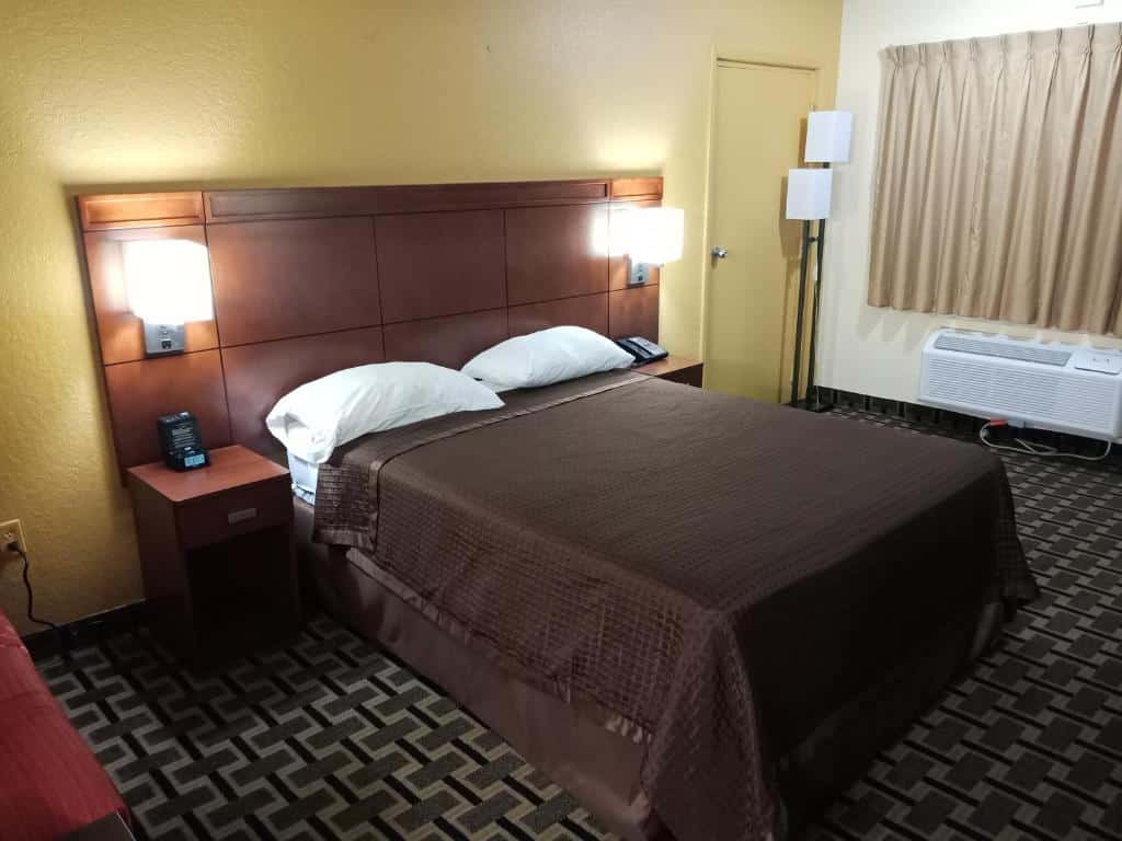 quarto com carpete, janela e aquecimento abaixo, com cama de casal e mesinha e luminária de ambos os lados, dá pra ver uma parte de um sofá na outra parede, no Budget Inn Of Orlando