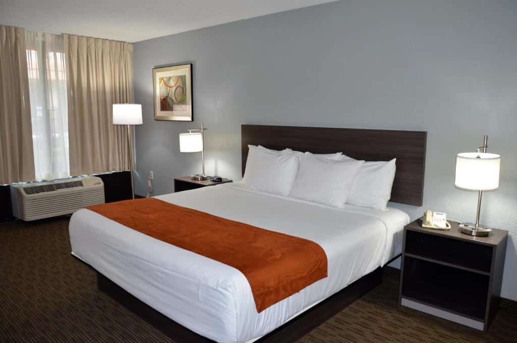 quarto do Days Inn & Suites by Wyndham Orlando Airport com cama de casal, luminárias de chão e mesinha ao lado da cama, janela grande com cortinas e o aquecedor abaixo