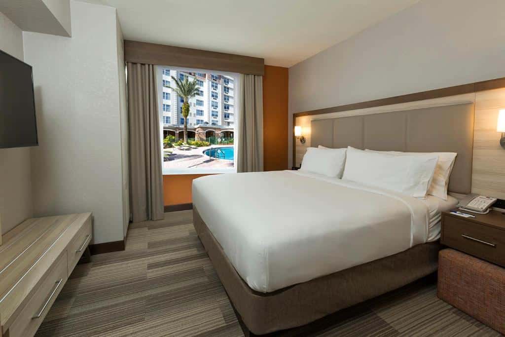 quarto do Holiday Inn Express & Suites S Lake Buena Vista, an IHG Hotel com janela com vista para a piscina, cama de casal com luminária e mesinha em ambos os lados, com tv à frente e uma cômoda