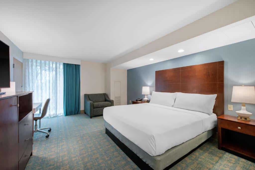 quarto do Holiday Inn Orlando – Disney Springs com cama grande, mesinha e luminária de ambos os lados, poltrona, mesa com cadeira e tv à frente e janela com cortinas escuras