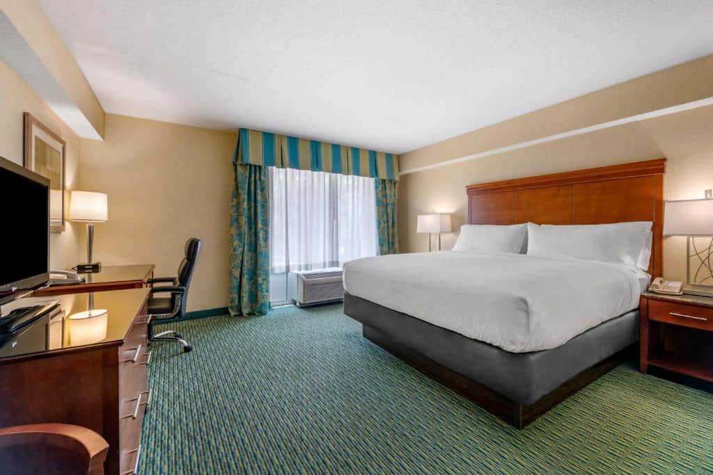 quarto amplo do Holiday Inn Resort Orlando com cômoda e tv e cadeira em frente à uma cama de casal com mesinha e luminária de ambos os lados, há aquecedor abaixo de uma janela com cortinas listradas verdes, que combinam com o carpete