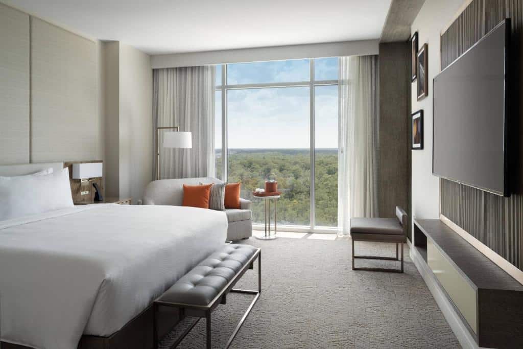 quarto do JW Marriott Orlando Bonnet Creek Resort & Spa com janela grande com vista, tv em frente à uma cama de casal com mesinha e luminária de ambos os lados, e um sofá, as cores são neutras e com detalhes em laranja